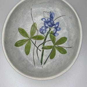 May include: A round, light gray ceramic bowl with a hand-painted bluebonnet floral design. The bowl features a central cluster of blue flowers, green leaves, and thin brown stems. The bowl's rim is slightly raised.