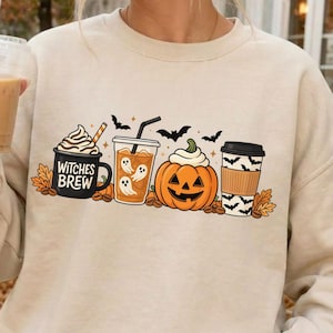 Op de afbeelding: Een beige sweatshirt met lange mouwen en een grafische print van een mok met de tekst Witches Brew, een glas met spookillustraties, een pompoen en een koffiebeker, omringd door herfstbladeren en kleine vliegende vleermuizen.