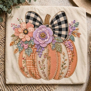 May include: A cream-colored t-shirt featuring a large embroidered pumpkin design. The pumpkin is composed of orange gingham and solid fabric panels, decorated with a black and white buffalo plaid bow and floral fabric appliques in pink and purple.