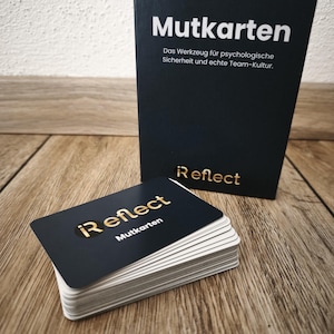 Può includere: Un mazzo di carte nere con scritte dorate, etichettate "i Reflect Mutkarten", è appoggiato su una superficie di legno accanto a una scatola nera con lo stesso testo. La scatola contiene anche il testo "Das Werkzeug für psychologische Sicherheit und echte Team-Kultur."