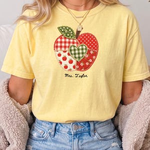 以下が含まれることがあります： 淡い黄色の半袖Tシャツ。赤と緑の様々な柄の布地をパッチワークしたリンゴの大きなグラフィックが特徴です。リンゴの中央には緑色のギンガムチェックのハートがあしらわれ、その下には黒い文字でMrs. Taylorとプリントされています。