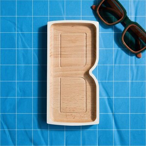 Puede incluir: Una bandeja de madera clara con forma de letra B, acabado liso y borde exterior blanco, situada junto a unas gafas de sol de montura oscura con cristales marrones. La bandeja presenta dos secciones rectangulares hundidas.