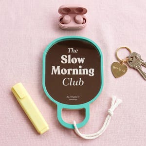 Può includere: Uno specchio con bordo verde acqua e centro marrone con il testo bianco The Slow Morning Club e ALPHABET home design. Sono inclusi auricolari wireless rosa in una custodia, un evidenziatore giallo e un portachiavi dorato con un cuore BFF's <3.
