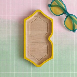 Puede incluir: Una bandeja de madera con un borde amarillo brillante en forma de letra B estilizada, que presenta dos compartimentos hexagonales empotrados. Junto a la bandeja hay unas gafas de sol con montura azul verdoso y lentes amarillas.