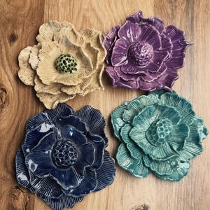 May include: Four ceramic flower wall sculptures in beige, purple, dark blue, and teal. Each piece features layered, textured petals and a central, raised, dotted seed pod. The surfaces have a glossy finish with visible ridges on the petals.