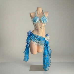 May include: A mermaid-themed costume on a mannequin. The outfit includes a seashell bra, a beaded necklace, and a blue skirt with floral details. The skirt has a sheer, flowing design with lace trim.