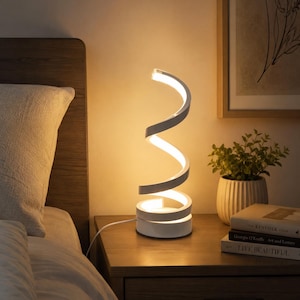 May include: A modern, white and silver spiral-shaped LED table lamp. The lamp is on a wooden nightstand next to a bed. The lamp's design is a continuous, twisting form, emitting a warm, inviting glow. The lamp is plugged into an electrical outlet.