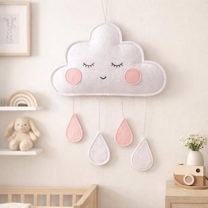May include: A white felt cloud-shaped wall hanging with a smiling face, pink cheeks, and closed eyes. Below the cloud hang pink and white teardrop shapes. This nursery decor item is designed to hang from a string.