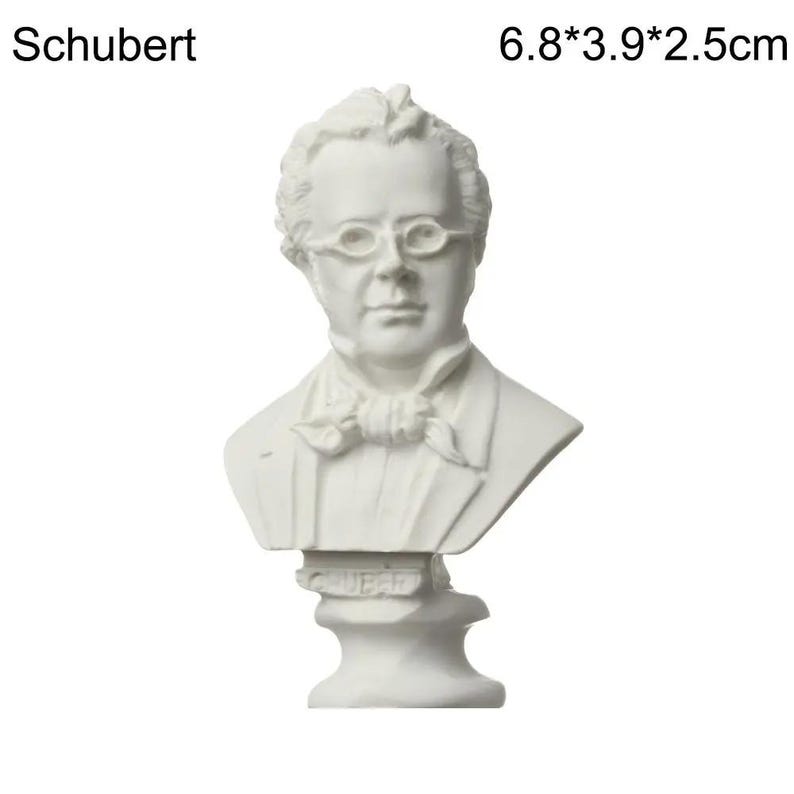 May include: A white bust of a man wearing glasses and a cravat. The bust is labeled "Schubert" and measures 6.8 x 3.9 x 2.5 cm. The sculpture is a classical style bust.