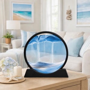 May include: A round, black-framed sand art piece with a blue and white sand design. The sand is suspended in a clear liquid, creating a dynamic, flowing landscape. The piece sits on a black base, perfect for a tabletop or shelf.