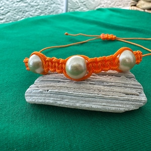 Op de afbeelding: Een oranje macramé armband met drie grote, ronde, crèmekleurige kralen. De armband wordt getoond op een stuk lichtgrijze steen tegen een groene achtergrond. De verstelbare sluiting is ook oranje.