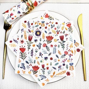 May include: A white plate setting with floral-patterned napkins, a rolled napkin with a gold ring, and gold cutlery. The napkins feature a colourful floral design with red, blue, yellow, and purple flowers and leaves.