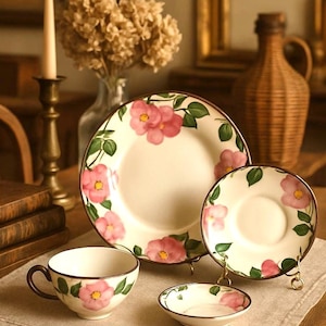 May include: A ceramic dinnerware set featuring a white base with hand-painted pink flowers and green leaves. The set includes a large dinner plate, a teacup with a dark handle, a saucer, and a small bowl, all finished with a thin brown rim.