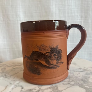 May include: A brown ceramic mug with a dark brown rim and handle. The mug features a detailed black illustration of a running hare. The mug is a rustic style and is suitable for hot or cold beverages.