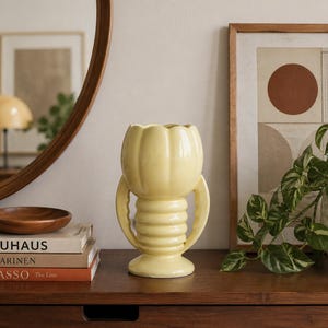 May include: A pale yellow ceramic vase with two handles and a scalloped rim. The vase is on a dark wooden surface, next to a stack of books and a decorative bowl. A framed print and a potted plant are also visible.