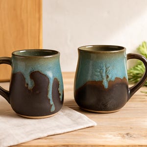May include: Two ceramic mugs featuring a two-tone glaze with a dark brown base and a turquoise blue top. The blue glaze drips down over the brown surface in a vertical pattern. Each mug has a curved handle and a smooth, rounded body shape.