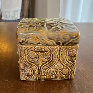 May include: A square ceramic box with a lid, featuring a glossy, mottled mustard-yellow and brown glaze. The exterior surfaces are decorated with a raised, intricate, symmetrical floral and scrollwork pattern that covers the entire box.