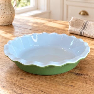 May include: A round ceramic pie dish featuring a smooth, light blue interior and a matte sage green exterior. The dish has a distinctively scalloped, fluted edge design around the entire rim, providing a decorative finish to the circular baking vessel.