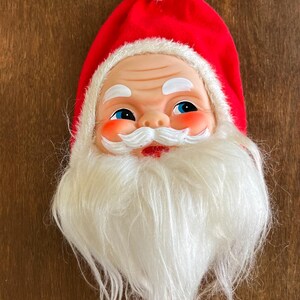 May include: A Santa Claus ornament featuring a molded plastic face with rosy cheeks, blue eyes, and a white mustache. The figure wears a red fabric hat with a white pom-pom and a white faux fur trim. A long white synthetic beard hangs below.