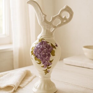 May include: A cream-colored ceramic pitcher featuring a hand-painted cluster of purple grapes with green leaves on the bulbous body. The vessel has a pedestal base, a narrow neck, and an ornate, openwork handle extending from the top rim.