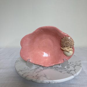 May include: A pink ceramic bowl with a scalloped edge, featuring a seashell accent. The bowl is displayed on a marble-patterned stand, creating a coastal or nautical theme. The bowl's dimensions are not visible.