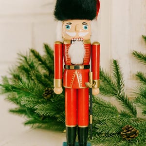 Puede incluir: Una figura de cascanueces de madera con un sombrero alto de piel negra, un uniforme rojo brillante con detalles dorados y botas negras. La figura tiene una barba de piel sintética blanca, rasgos faciales pintados y una base rectangular azul claro.