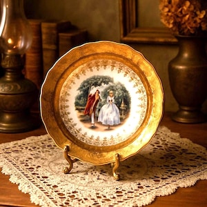 May include: A round decorative plate with a scalloped edge and a wide, textured gold rim. The center features a circular illustration of two figures in historical attire standing in a park setting, surrounded by a detailed gold filigree border.