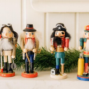 May include: Four wooden nutcracker figurines standing in a row. The first wears a pilot hat and goggles, the second a black hat, the third a pirate hat with an eyepatch, and the fourth a colourful cap. Each figure features a white beard and uniform.