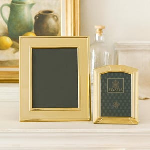 May include: Two gold-toned metal picture frames. The larger frame is rectangular with a smooth finish. The smaller frame has a rounded top with ribbed detailing and contains a dark green insert with the text ELYSEES and a crest logo printed in gold.