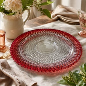 Op de afbeelding: Een heldere glazen serveerschaal met een rode rand en een textuur van diamantpatroon. De schaal staat op een wit tafelkleed, met een vaas met bloemen en een glas roze vloeistof op de achtergrond. De schaal heeft een diameter van ongeveer 30 cm.