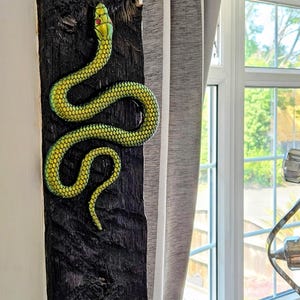 May include: A metallic green snake sculpture with a textured scale pattern and small red eyes, mounted vertically on a dark, rough-hewn wooden beam. The snake is depicted in a winding, S-shaped posture, showcasing a vibrant, reflective finish.