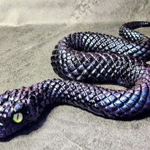 May include: A coiled snake figurine featuring a textured, diamond-patterned surface with a deep purple and black iridescent finish. The snake has a single visible eye with a bright yellow iris and a vertical black pupil, positioned on its head.