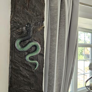 May include: A three-dimensional snake sculpture mounted on a dark, textured wooden beam. The snake features a black head and upper body that transitions into a vibrant, iridescent green and purple scaled pattern along its winding, S-shaped body.