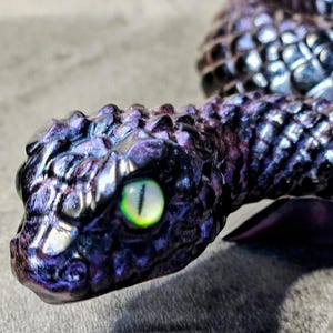 May include: A coiled snake figurine featuring a dark purple and black iridescent scale texture. The snake has a prominent, detailed head with a single visible eye that is bright yellow-green with a vertical black pupil, reflecting light.