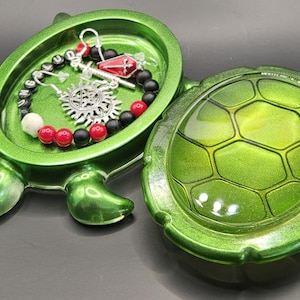 May include: A green turtle-shaped trinket box with a removable lid. Inside the open base rests a beaded bracelet featuring black and red beads, a silver pentagram charm, and a small red coffin charm with a silver cross, plus lettered beads.