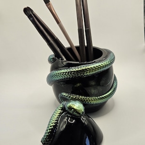 May include: A black cylindrical container with a glossy finish, featuring a sculpted iridescent green and blue snake coiled around the exterior. A matching black lid with a snake head rests in front, and four makeup brushes are inside the container.