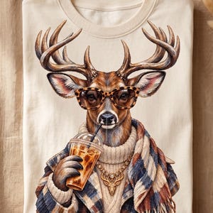 Könnte beinhalten: Ein cremefarbenes T-Shirt mit einer Grafik eines Hirsches, der eine Sonnenbrille mit Leopardenmuster, einen cremefarbenen Zopfmusterpullover, eine goldene Hirsch-Halskette und einen karierten Schal trägt und einen Eiskaffee im Plastikbecher hält.
