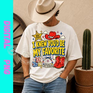 May include: A cream-colored t-shirt featuring a graphic print with a red cowboy hat, alphabet blocks, a beaded bracelet, and red cowboy boots. The text reads "BEST BUDS" and "I KNEW YOU'D BE MY FAVORITE" in bold yellow and blue lettering.