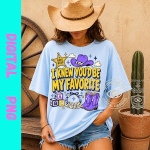May include: A light blue t-shirt featuring a graphic print with the text I KNEW YOU'D BE MY FAVORITE. The design includes a yellow sheriff star, a purple cowboy hat, alphabet blocks, a beaded bracelet, and purple cowboy boots with stars.