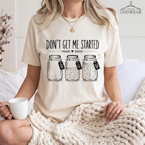 Pode incluir: Camiseta creme com a frase "DON'T GET ME STARTED" acima de três potes de vidro ilustrados. Os potes têm conteúdos e etiquetas variados. A camiseta é de estilo casual.