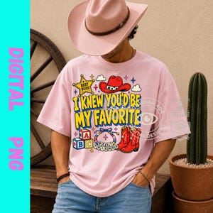May include: A light pink t-shirt featuring a graphic print with a yellow sheriff star labeled Best Buds, a red cowboy hat, and the text I KNEW YOU'D BE MY FAVORITE in yellow. Below are alphabet blocks, a beaded bracelet, and red cowboy boots.