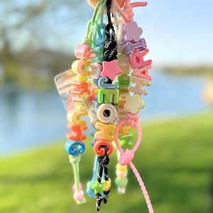 May include: A collection of colorful beaded bracelets spelling out "HOME" and "HAPPY". The bracelets feature various shapes like stars and flowers, with a mix of bright colors including pink, green, and yellow. The background is a blurred view of a green field and blue water.