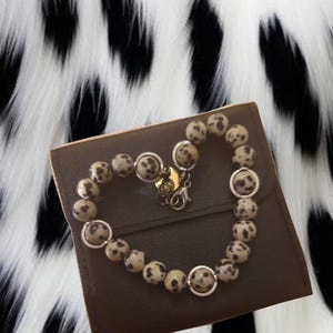 May include: A heart-shaped bracelet made of speckled, round beads, resting on a brown leather case. The bracelet is secured with a silver clasp. Matching earrings are visible at the top. The background is a black and white spotted pattern.