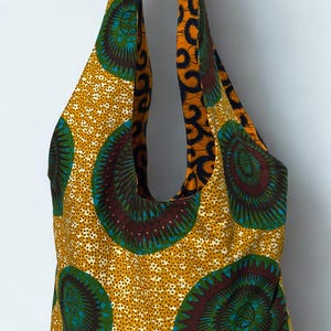 Pode incluir: Bolsa tote estampada com fundo amarelo mostarda e grandes desenhos circulares em verde-azulado, castanho e verde. As alças e o interior da bolsa apresentam um padrão de redemoinho laranja e azul marinho contrastante. A bolsa é de tecido.