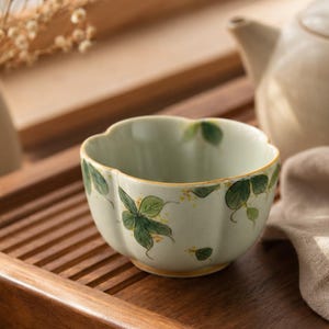 May include: A pale green ceramic tea bowl with a scalloped rim and a hand-painted botanical pattern featuring green leaves and small yellow flowers. The bowl has a smooth, rounded shape and a subtle yellow trim along the top edge of the rim.