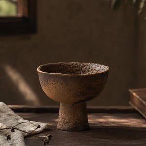 Può includere: Una ciotola in ceramica marrone strutturata con base a piedistallo. La ciotola ha una parte superiore ampia e aperta e una superficie ruvida e irregolare. Anche la base del piedistallo è strutturata. La ciotola è appoggiata su una superficie di legno scuro, con un panno bianco e una finestra sullo sfondo.