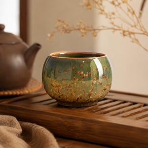 May include: A small, round ceramic tea cup with a green and brown speckled glaze. The cup has a golden rim and sits on a wooden tray. A brown teapot is partially visible in the background, suggesting a tea set.