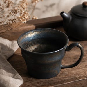 May include: A black ceramic teacup with a curved handle and a gold rim sits on a wooden surface. A black teapot is in the background. The teacup has a textured exterior and a wide opening.