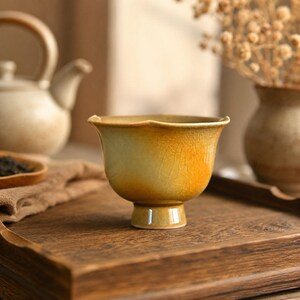May include: A ceramic tea cup featuring a yellow and light green gradient glaze with a visible crackle pattern. The cup has a wide, flared rim with a subtle scalloped edge and sits on a raised, cylindrical pedestal base, showcasing a smooth finish.