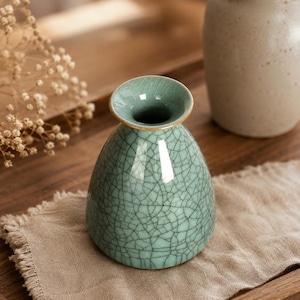 Può includere: Un piccolo vaso in ceramica a forma di campana con una smaltatura verde acqua chiaro e un evidente motivo a crepe scure. Il vaso presenta un bordo circolare ampio e svasato con un sottile profilo dorato, creando una silhouette liscia e tonda.